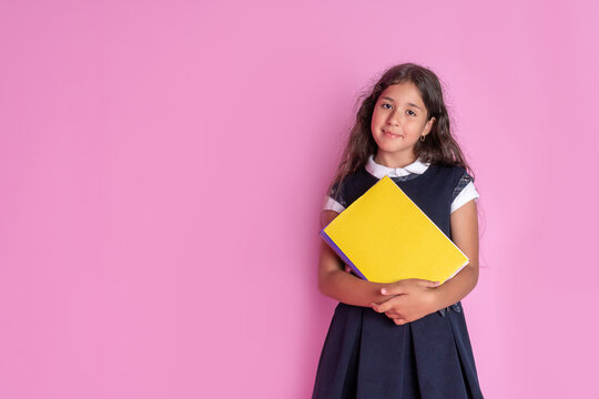 A charming girl with long curly dark hair in a school uniform with a book in her hands on a pink background. Studio photo. Back to school.