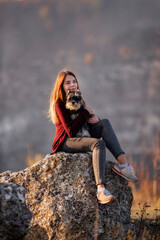 A beautiful girl with red hair holds a black Schnauzer in her arms. A model sits on a large stone, hugs a dog, against a background of autumn textures. Traveling with pets. Caring, love for smaller 