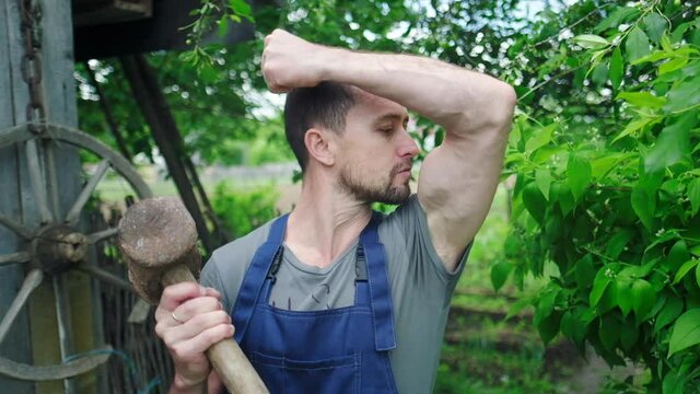 Funny worker in the garden. Young adult man stands in the summer garden with big hammer and kisses his own biceps
