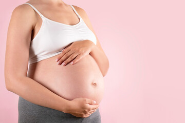 Close up of pregnant woman wearing supportive seamless maternity bra & grey yoga pants, arms on her belly. Female hands wrapped around big bare tummy. Child expectancy concept. Background, copy space