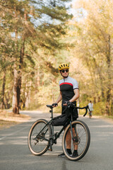 Cyclist in a sports outfit poses for a camera on the road in an autumn forest, stands with a bicycle rider and looks away with serious face. Concentrated young man with a bicycle relaxing on park