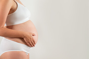 Close up of pregnant woman wearing supportive seamless maternity bra and maxi bottoms, arms on her belly. Female hands wrapped around big bare tummy. Child expectancy concept. Background, copy space