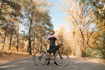 Young man in sportswear standing with bicycle on background of autumn landscape and looking away. Professional cyclist standing on road in autumn forest outside city.