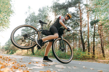 Fototapeta na wymiar Male cyclist in sportswear and helmet doing trick on bicycle on road in autumn forest at sunset.Professional valeopedist effectively lifts the rear wheel of a bicycle while training in an autumn park
