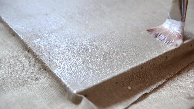 Paintbrush applying gesso on raw linen canvas, oil painting priming close up