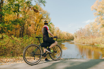 Professional cyclist in sports outfit stands on the shore of a lake in the park on a beautiful autumn landscape. Cyclist sits on a bicycle bike and relaxes looking at the beautiful autumn scenery.