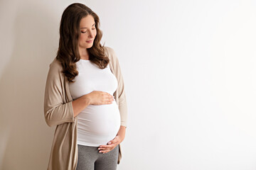 Portrait shot of young beautiful woman on third trimester of pregnancy. Close up of pregnant female with arms on her round belly. Expecting a child concept. Background, copy space. - obrazy, fototapety, plakaty