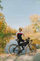 Professional cyclist standing with bicycle against the background of beautiful autumn landscape with river and forest. Cycling sportswear and helmet rest on a walk in the beautiful park.