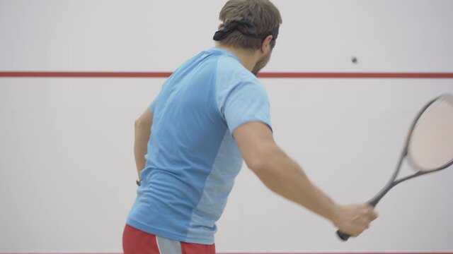 Middle shot of brunette bearded Caucasian man hitting squash ball against the wall. Side view portrait of confident mid-adult sportsman using racket to play sport game in gym indoors.