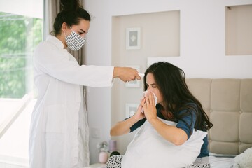 Female sneezing and covering her face with a tissue while having her fever checked by a nurse