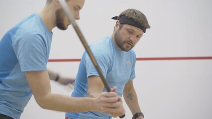 Side view of young coach correcting posture and gripping technique of bearded tattooed sportsman playing squash in gym. Confident Caucasian men training indoors.