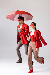 full length view of trendy couple in red blazers and sunglasses running with umbrella on grey