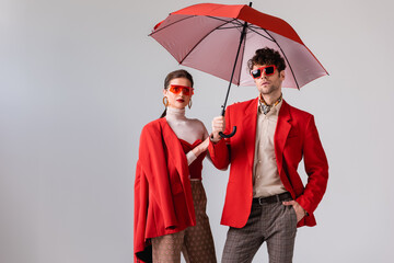young, stylish couple in red blazers and sunglasses posing with umbrella isolated on grey