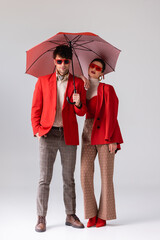 full length view of young trendy couple in red blazers posing with umbrella on grey