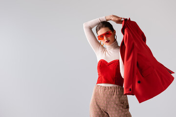 fashionable girl in sunglasses holding red blazer while looking at camera isolated on grey