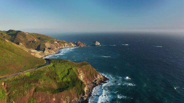 4K nature travel and outdoor adventure. Cinematic aerial view of famous California highway N1 running along scenic rocky Pacific Ocean coast. Clear blue sky above beautiful seascape at sunset, USA