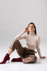 attractive, fashionable woman looking at camera while sitting on floor on grey background