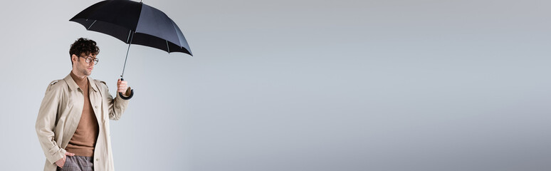 panoramic crop of fashionable man standing under umbrella with hand in pocket on grey © LIGHTFIELD STUDIOS
