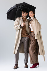 full length view of young couple in fashionable autumn clothes posing under umbrella on grey