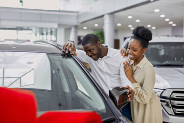 handsome dark-skinned man pleases his wife in dealership, he give car as a present, happy woman and man in love - obrazy, fototapety, plakaty