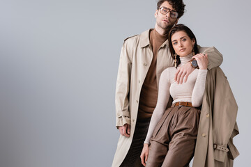 trendy man in trench coat hugging fashionable woman while looking at camera isolated on grey