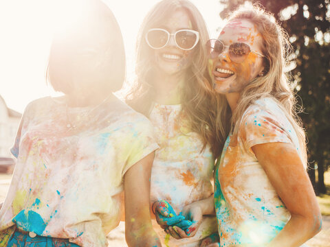 Three happy beautiful girls making party at Holi colors festival in summer time.Young smiling women friends having fun after music event at sunset. Positive models going crazy in sunglasses at sunset