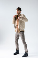 Fototapeta premium full length view of stylish man touching jacket while looking at camera on grey