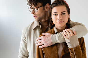 stylish man in eyeglasses hugging stylish woman looking at camera on grey