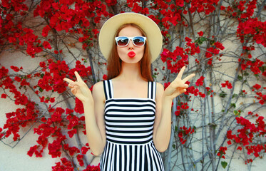 Attractive woman blowing red lips sending sweet air kiss wearing a summer straw hat over red flowers background
