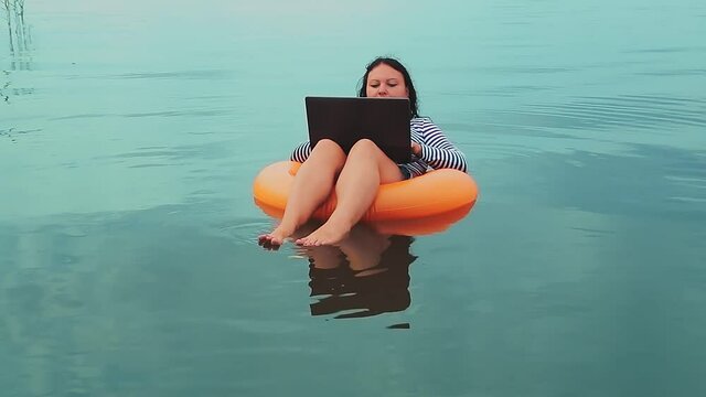 A young woman freelancer in an inflatable ring swims near the sea in the morning and works in a laptop.