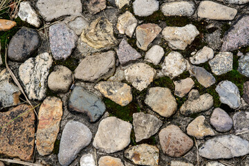 A background of small stones lying close to each other, of different shapes, colors and textures, embedded in the ground and overgrown with grass and moss in between.