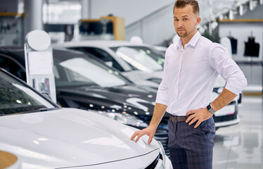 portrait of happy caucasian client man buying new car in car store. luxurious white car is on exhibition in dealership