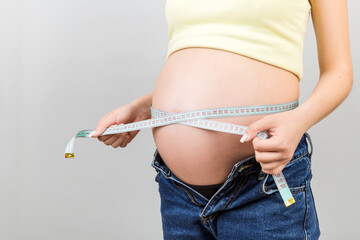 Close up of a pregnant woman in opened jeans measuring her belly with a tape measure at colorful background with copy space. entimeter measurement