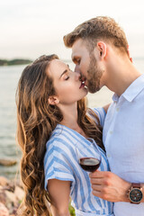 bearded man kissing girlfriend and holding glass with red wine near lake