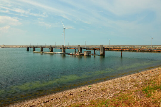 Brown pebble beach and deep blue water, a long pier with wooden pillars, wind turbine and dam in the background. Netherlands, Vrowenpolder.