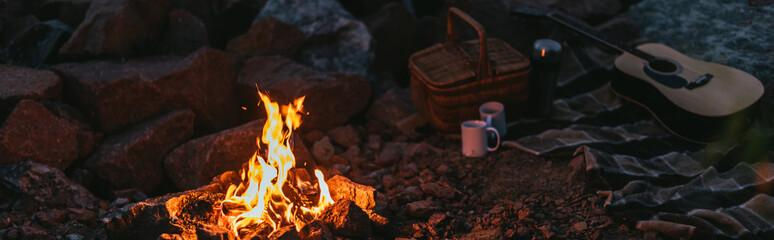 panoramic crop of burning bonfire near plaid blanket, wicker basket, cups and acoustic guitar