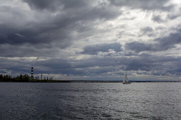 Fototapeta na wymiar White sailboat and stripped lighthouse at place where Daugava river flows into the Baltic sea, under dramatic sky.
