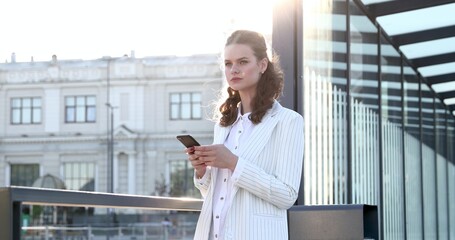 Attractive woman in formal clothing standing on station and searching information about bus traffic on cellphone. Young girl with brown hair using mobile application outdoors.