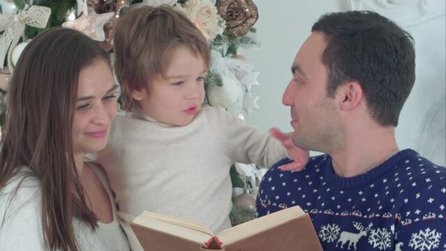 Happy father reading about Santa beard, while mother holding their playful son in front of the Christmas tree