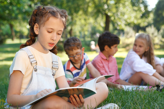 Young girl reading a book at the park, her friends relaxing on the grass on background