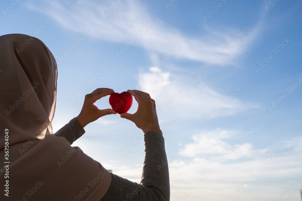 Wall mural silhouette of muslim woman in head scarf with red heart on blue sunset sky background. concept