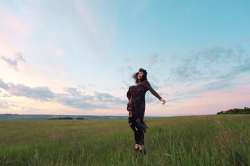 Fototapeta premium A young woman of 25-30 years old with curly red hair, in a green dress and a diadem with feathers, is spinning and dancing against the background of beautiful nature at dawn. 