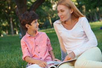 Cute happy little boy smiling at his female teacher, sitting with a book in the grass