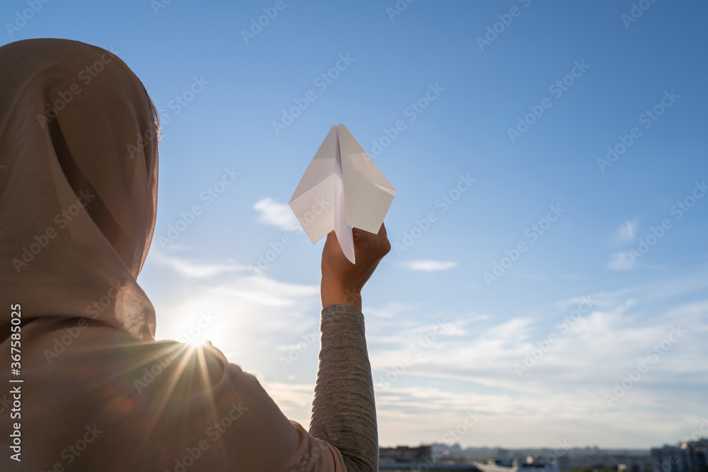 Wall mural silhouette of muslim woman in head scarf launching a paper airplane on blue sunset sky background. t