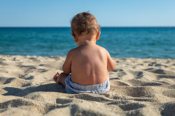 Authentic back view shot of a toddler baby boy is sitting and playing with a sand on a sandy beach during family vacation holiday at sea.