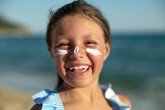 Happy Little Girl Is Smiling In Camera With A Protective Sunscreen Or Sunblock Lotion On Her Face On A Seaside Sandy Beach During Family Holidays Vacation.
