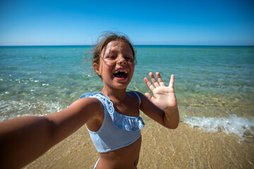 A happy carefree little girl is having fun to make a selfie or technology video call to parents or friends on a beach with seaside in a sunny day during family vacation holidays.
