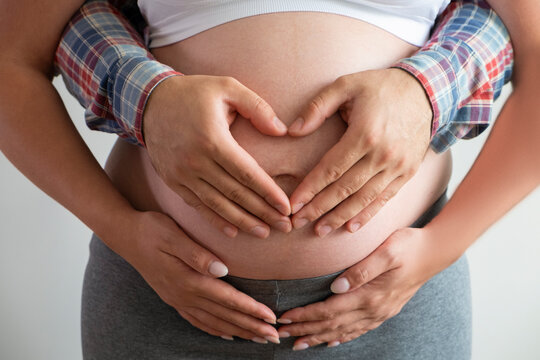 Close up shot of young man hugging his pregnant wife from behind, forming a heart shape with his hands over isolated white background. Prenatal period concept. Young family expecting a new child.