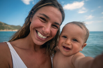 Fototapeta na wymiar Authentic close up shot of happy carefree smiling neo mother and her newborn baby making selfie or video call to father or relatives on a beach with seaside in a sunny day. 