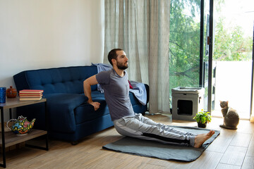 A young man doing dips workout at home using a sofa during the morning in pandemic lockdown. His cat watches